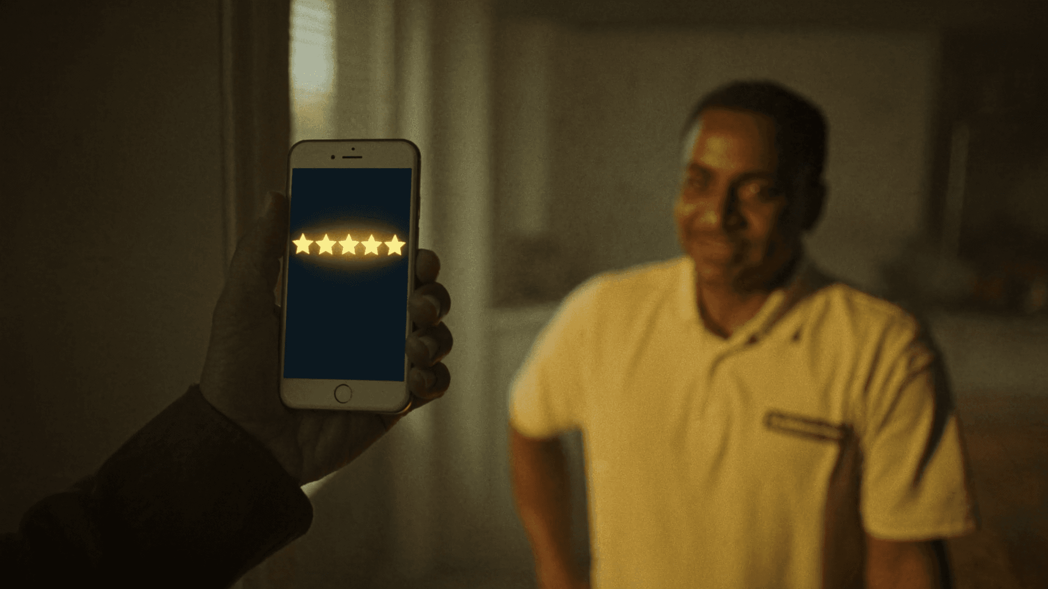 A homeowner's hand holding a smartphone showing five gold rating stars in a sunlit kitchen, a home-service contractor smiling out of focus behind