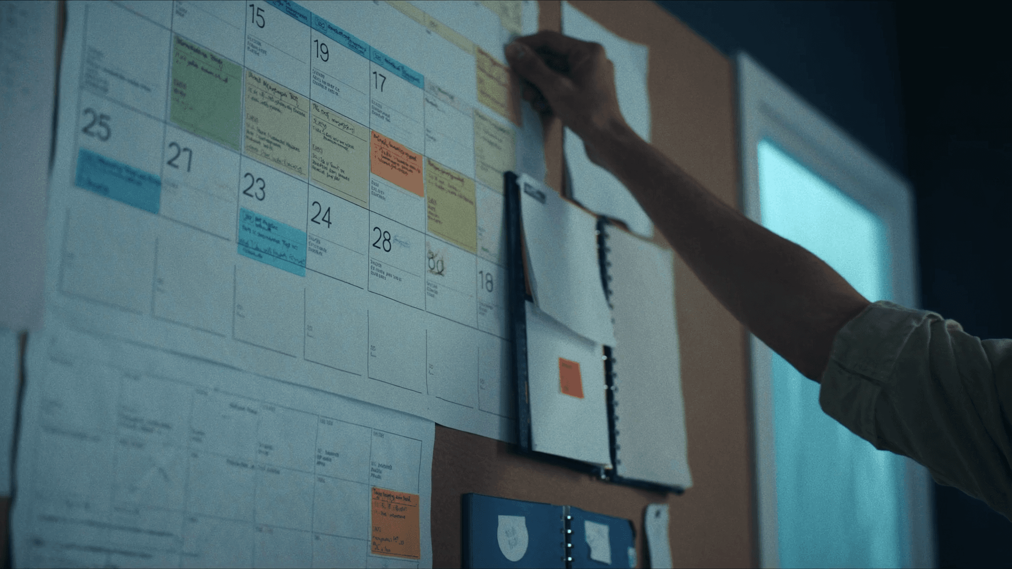 A busy wall calendar and desk planner in a tidy home office, every day filled with color-coded appointment blocks, a contractor's hand pinning a note in cool morning light